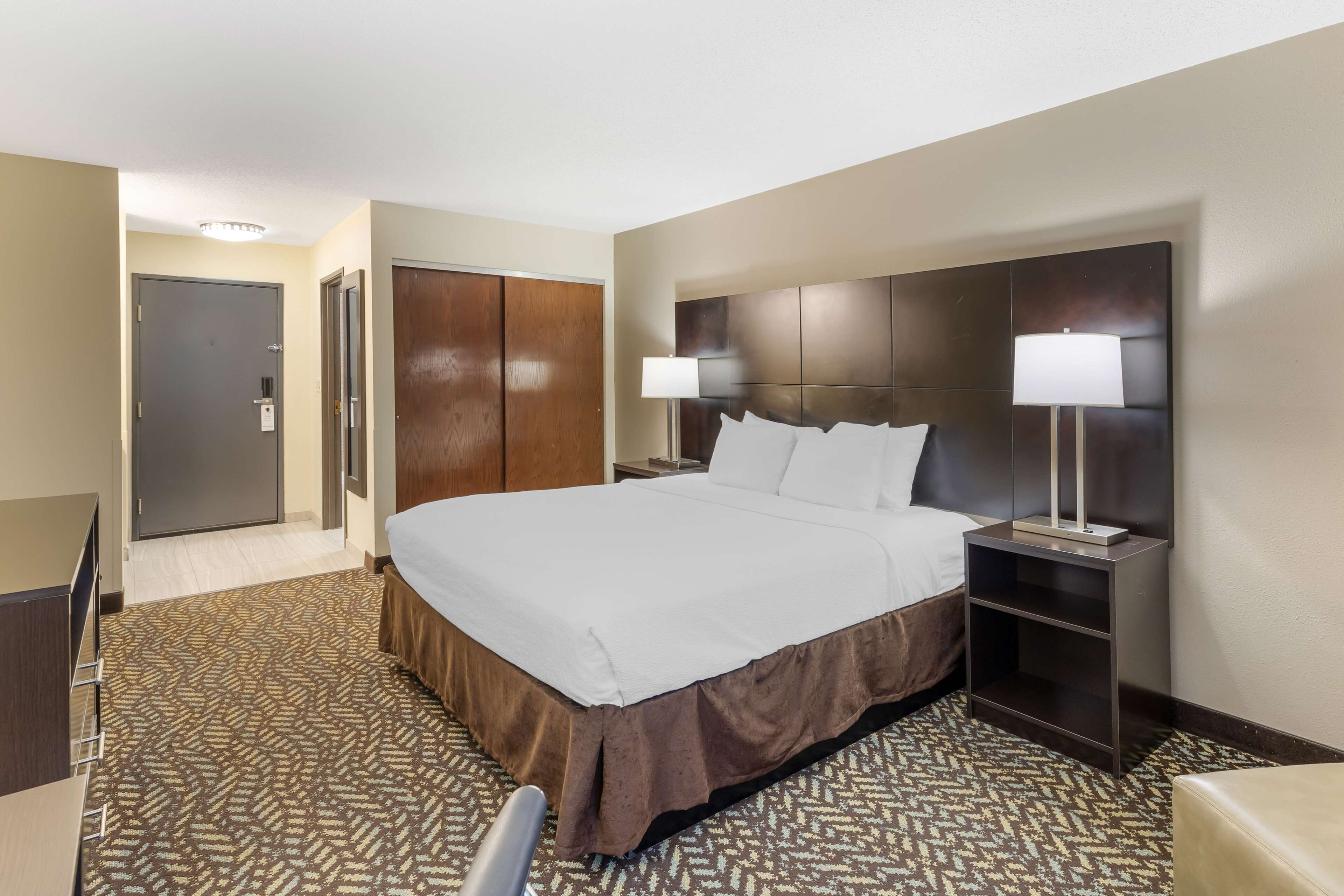 A hotel room with a large bed, a desk, a wardrobe, and a nightstand with a lamp.