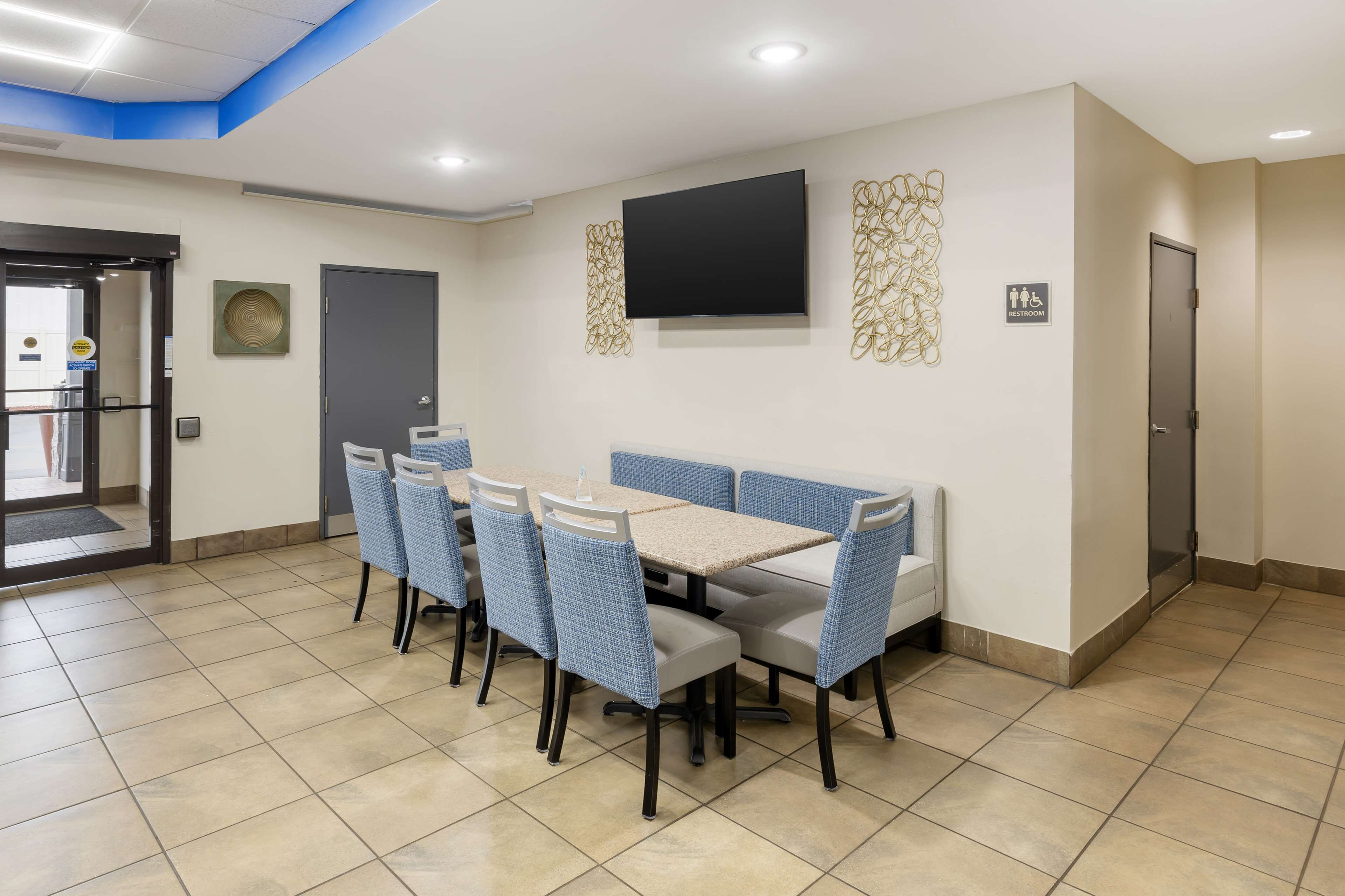 Une chambre d’hôtel dotée d’un coin repas avec une longue table et des chaises bleues, un téléviseur à écran plat fixé au mur, et un panneau indiquant l’emplacement des toilettes.