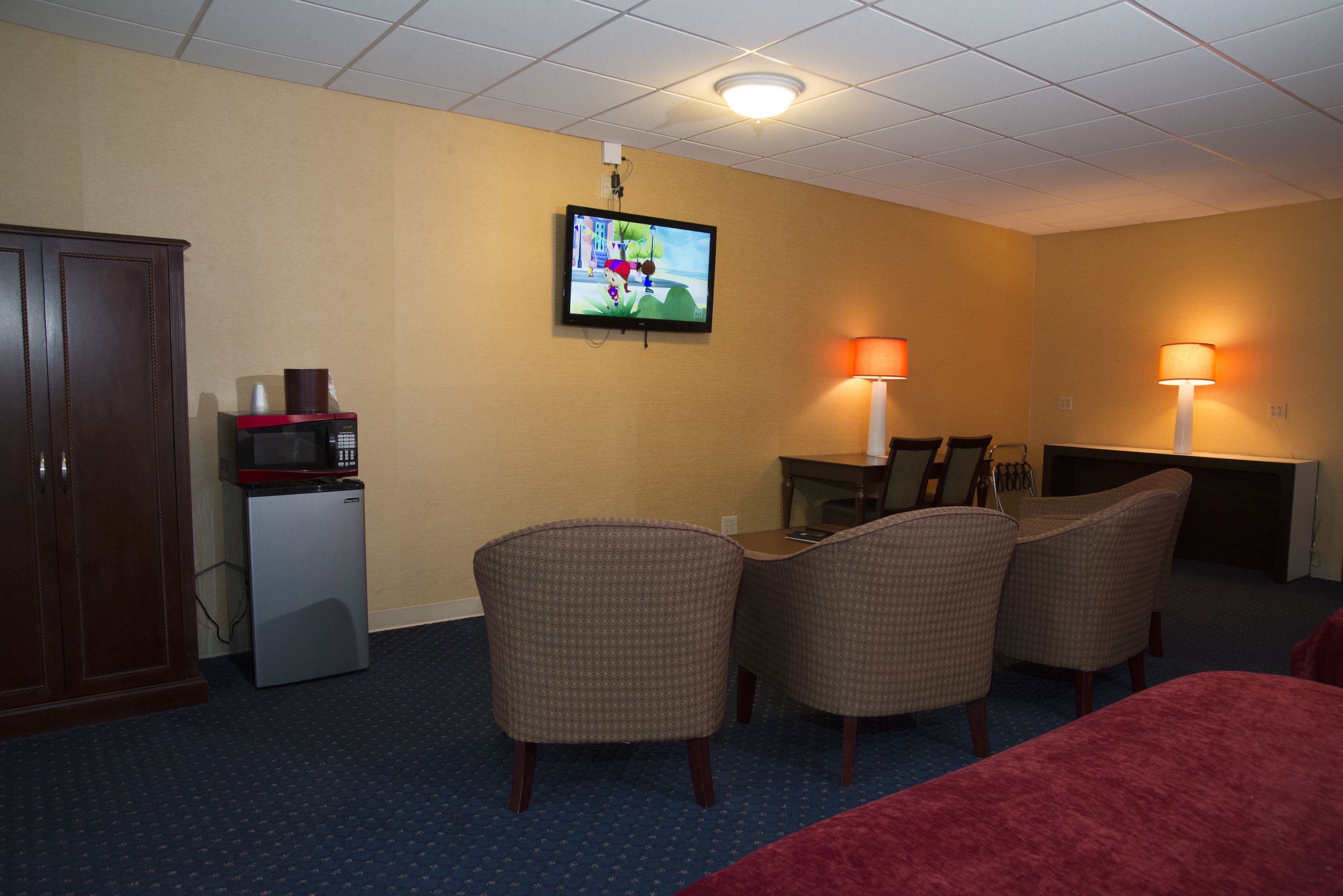 A hotel room with a television mounted on the wall, a desk with a lamp, and two chairs.