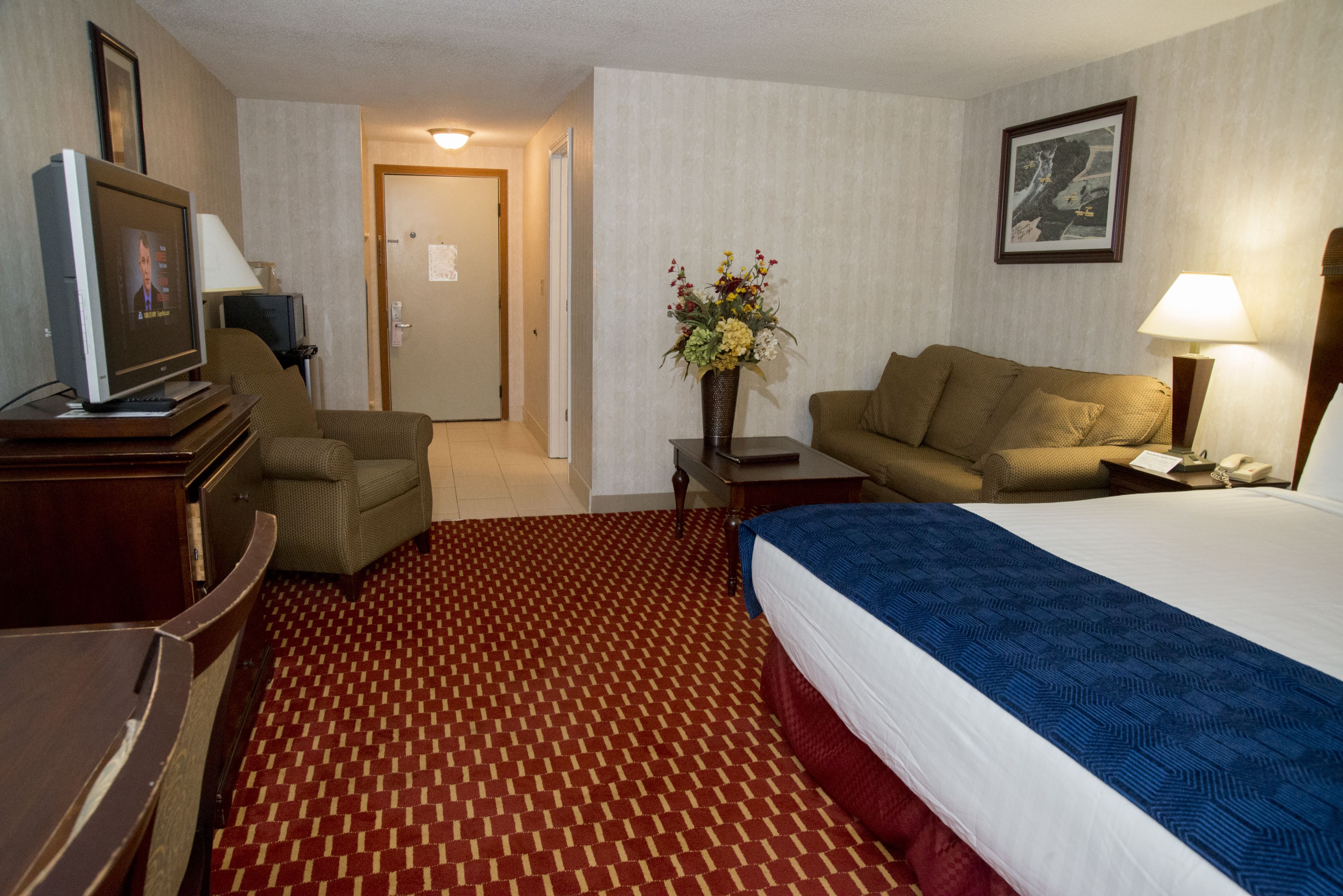 A hotel room with a large bed, two armchairs, a desk with a computer, a TV, and a vase with flowers.