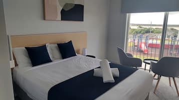 A hotel room with a large bed, a headboard, two pillows, a blanket, a bedside table, a lamp, a chair, and a view of a playground through a window.