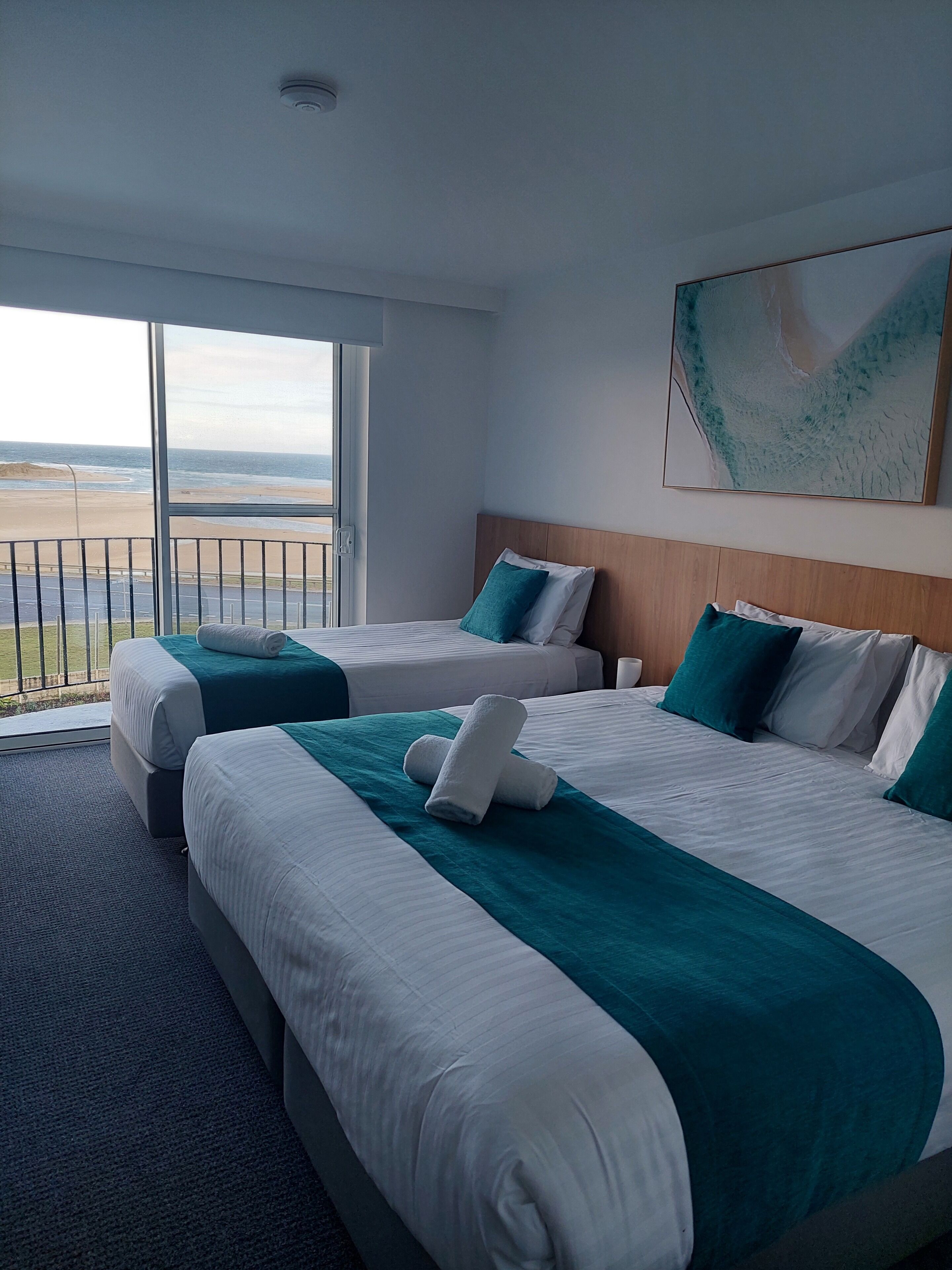 A hotel room with two beds, a large window with a view of the beach, and a painting on the wall.