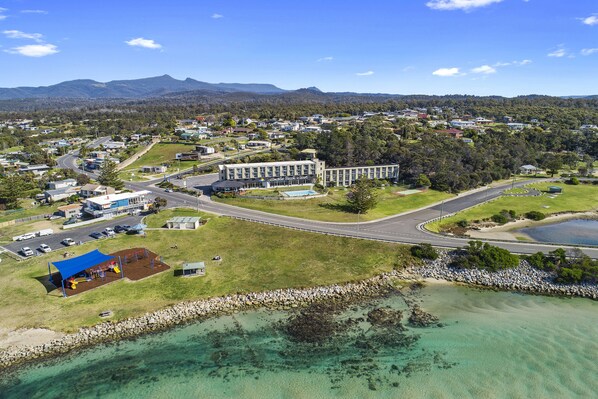 An aerial view of a coastal town with a prominent building, a playground, and clear turquoise waters.