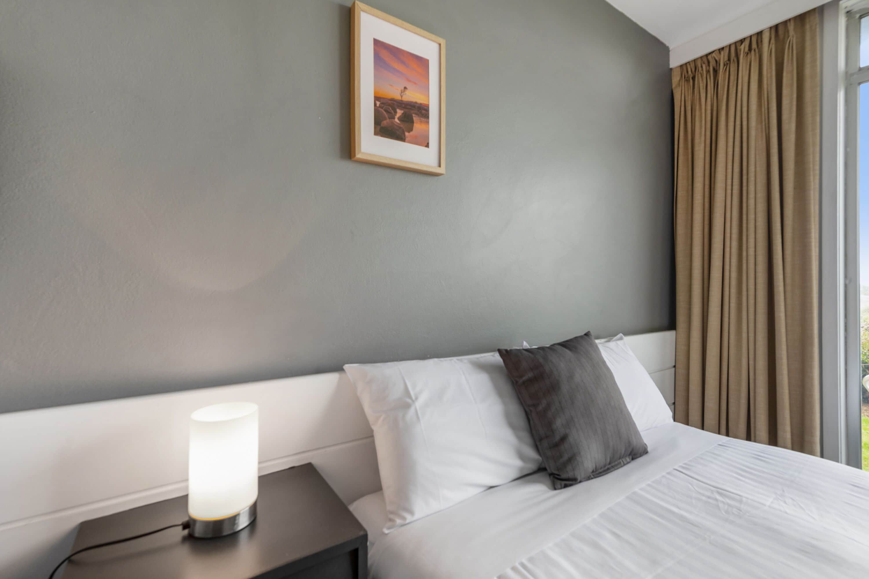 Una habitación de hotel con cama, lámpara de noche, un cuadro en la pared y ventana con cortinas.