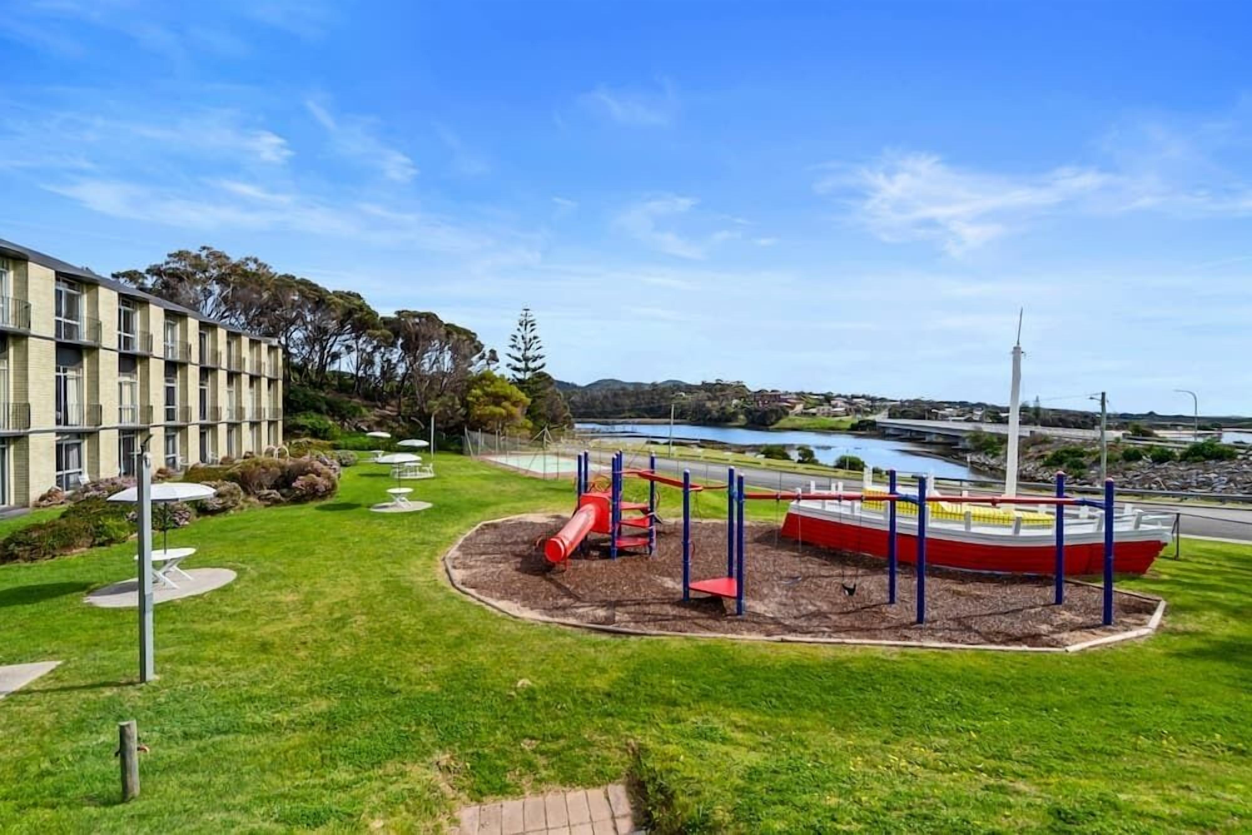 A playground with slides and climbing structures, surrounded by a well-maintained lawn and a building with multiple windows.