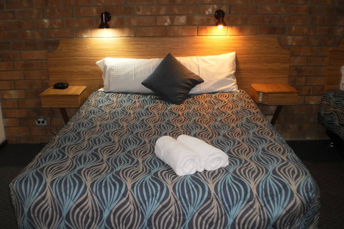 A neatly made bed with a patterned bedspread, two pillows, and a rolled white towel. There are two bedside tables with lamps, and a brick wall in the background.