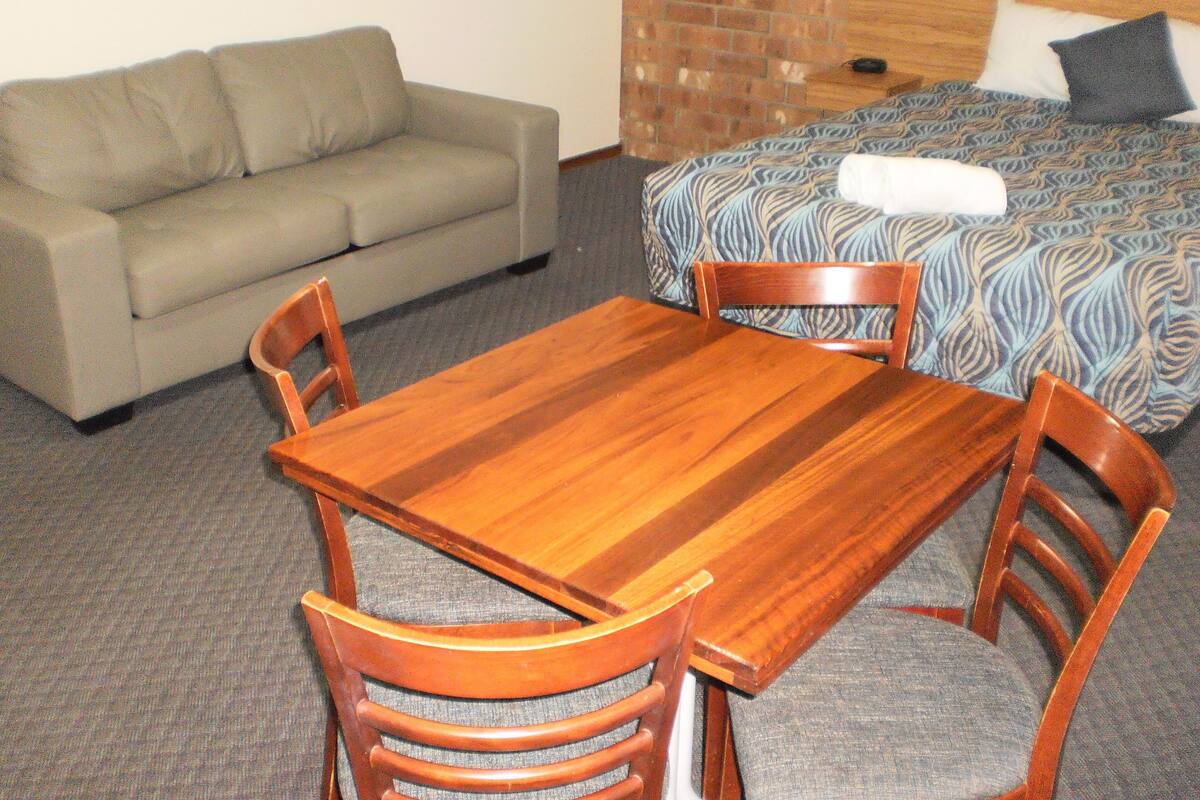 A room with a wooden dining table, four chairs, a sofa, and a bed with patterned bedding.