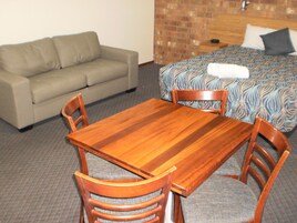 A room with a wooden dining table, four chairs, a sofa, and a bed with patterned bedding.