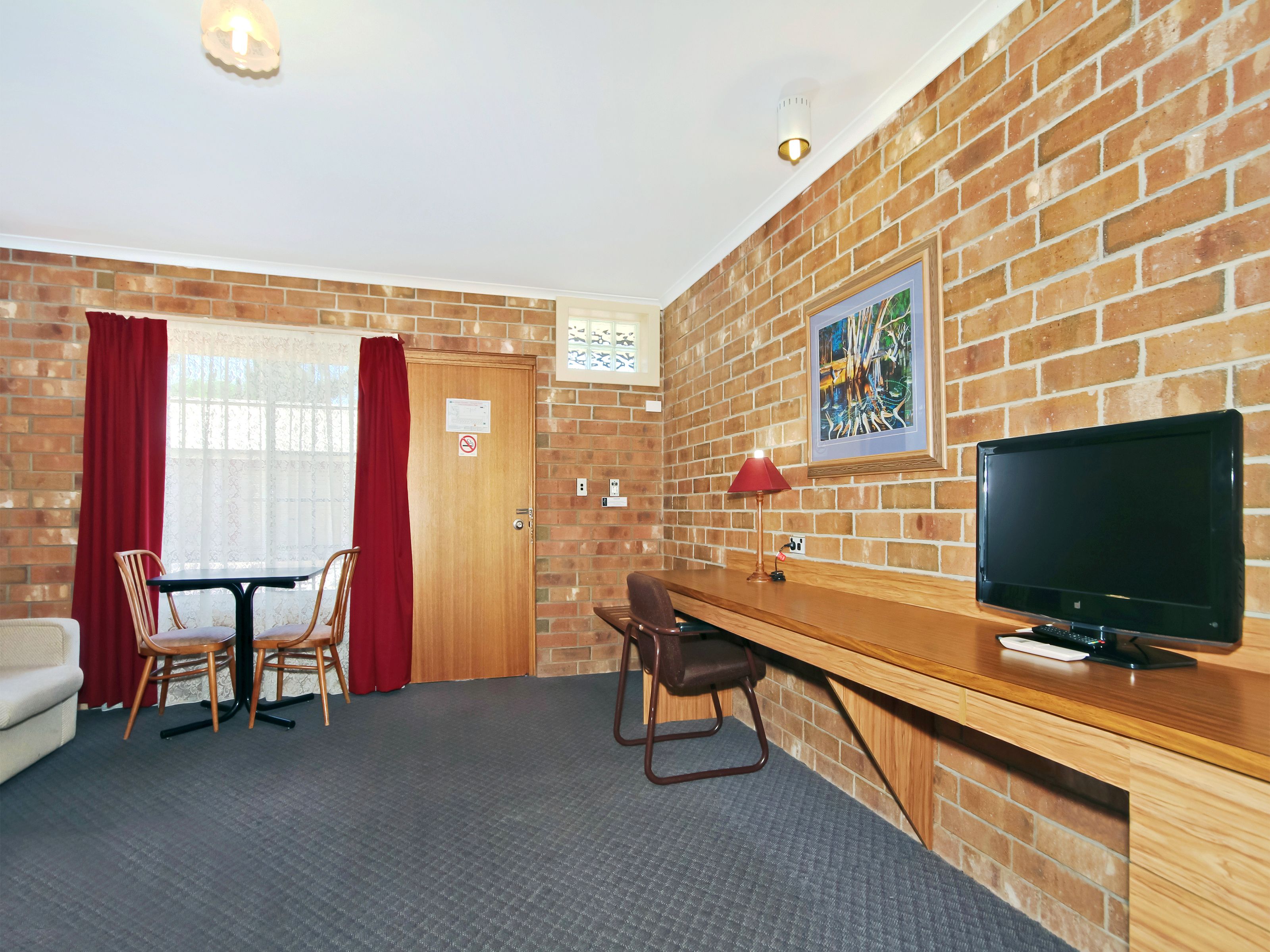 A room with a brick wall, a wooden desk with a computer and lamp, a TV, a chair, a table, and a couch.