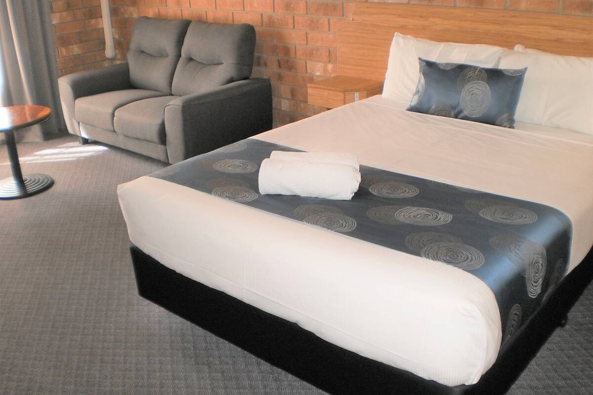 A hotel room with a large bed, a grey armchair, and a small table.