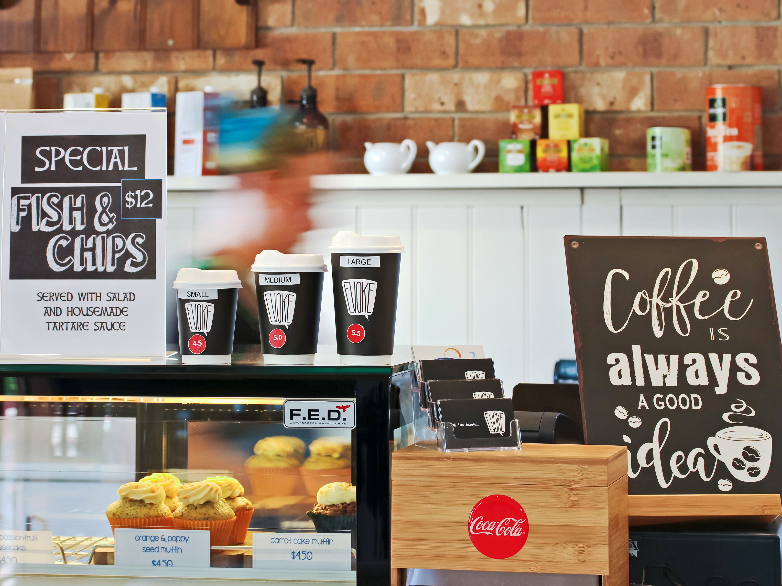 L’intérieur d’un café présente un panneau indiquant une offre spéciale de poisson-frites à 12 dollars, différentes tailles de tasses de café, et un autre panneau sur lequel on peut lire : « Un café, c’est toujours une bonne idée ».