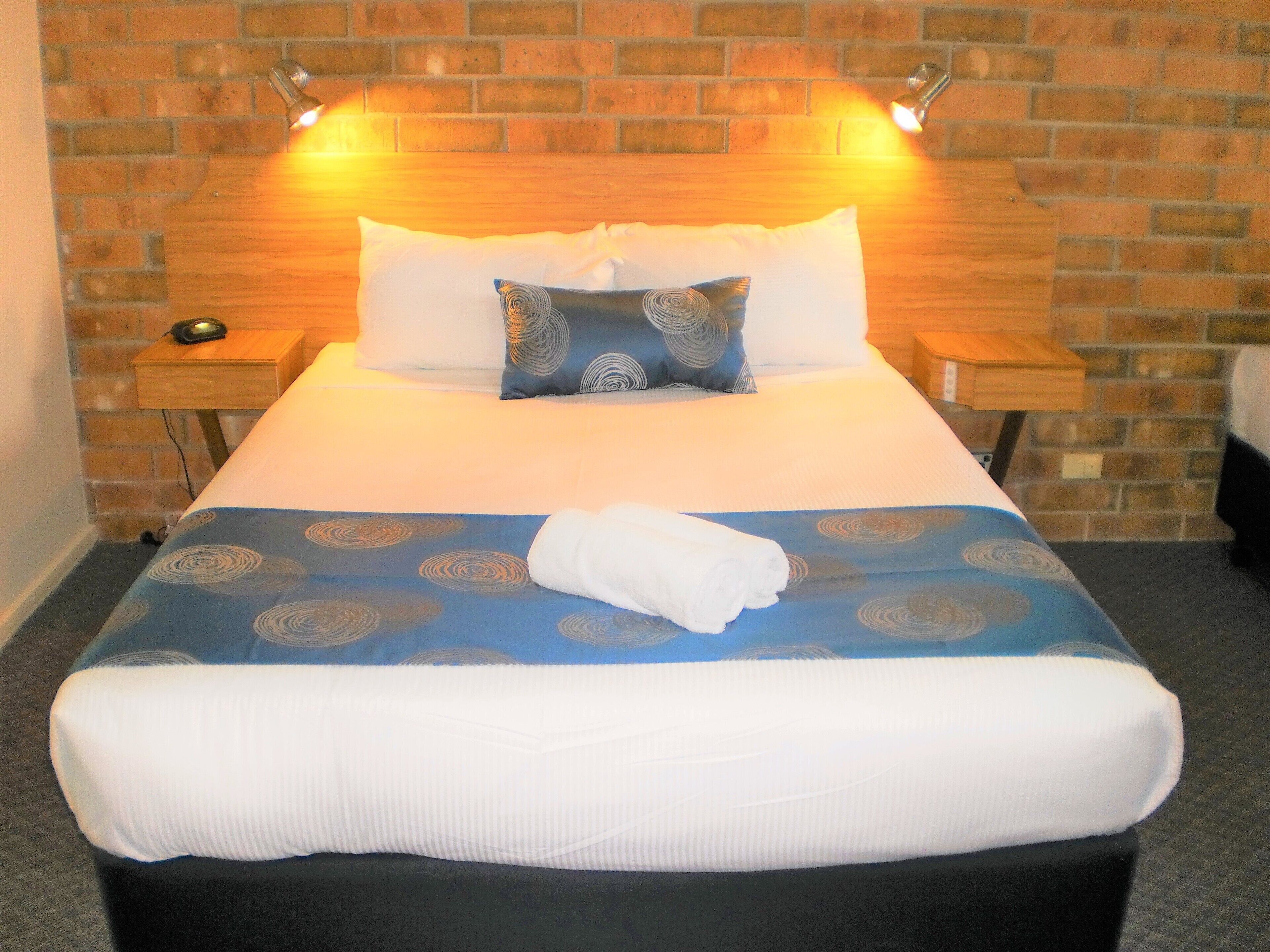 A neatly made bed with a blue and white patterned bedspread, two pillows, and a folded white towel. There are two bedside tables with lamps, and a brick wall in the background.