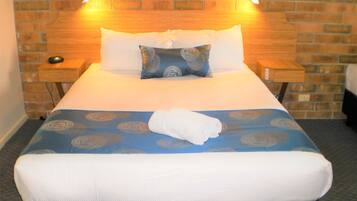 A neatly made bed with a blue and white patterned bedspread, two pillows, and a folded white towel. There are two bedside tables with lamps, and a brick wall in the background.