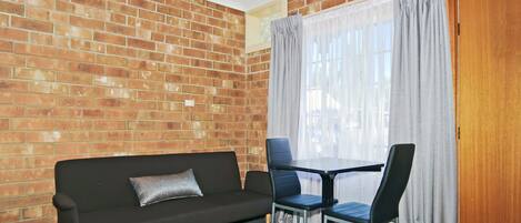 A room with a brick wall, a black sofa with a cushion, two black chairs, a small table, and a wooden door.