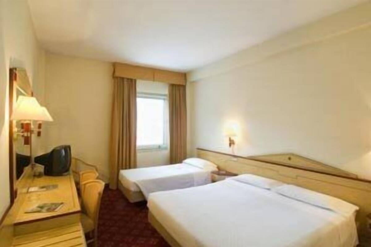 Une chambre d’hôtel avec deux lits, un bureau avec une lampe, une chaise et une fenêtre avec des rideaux.