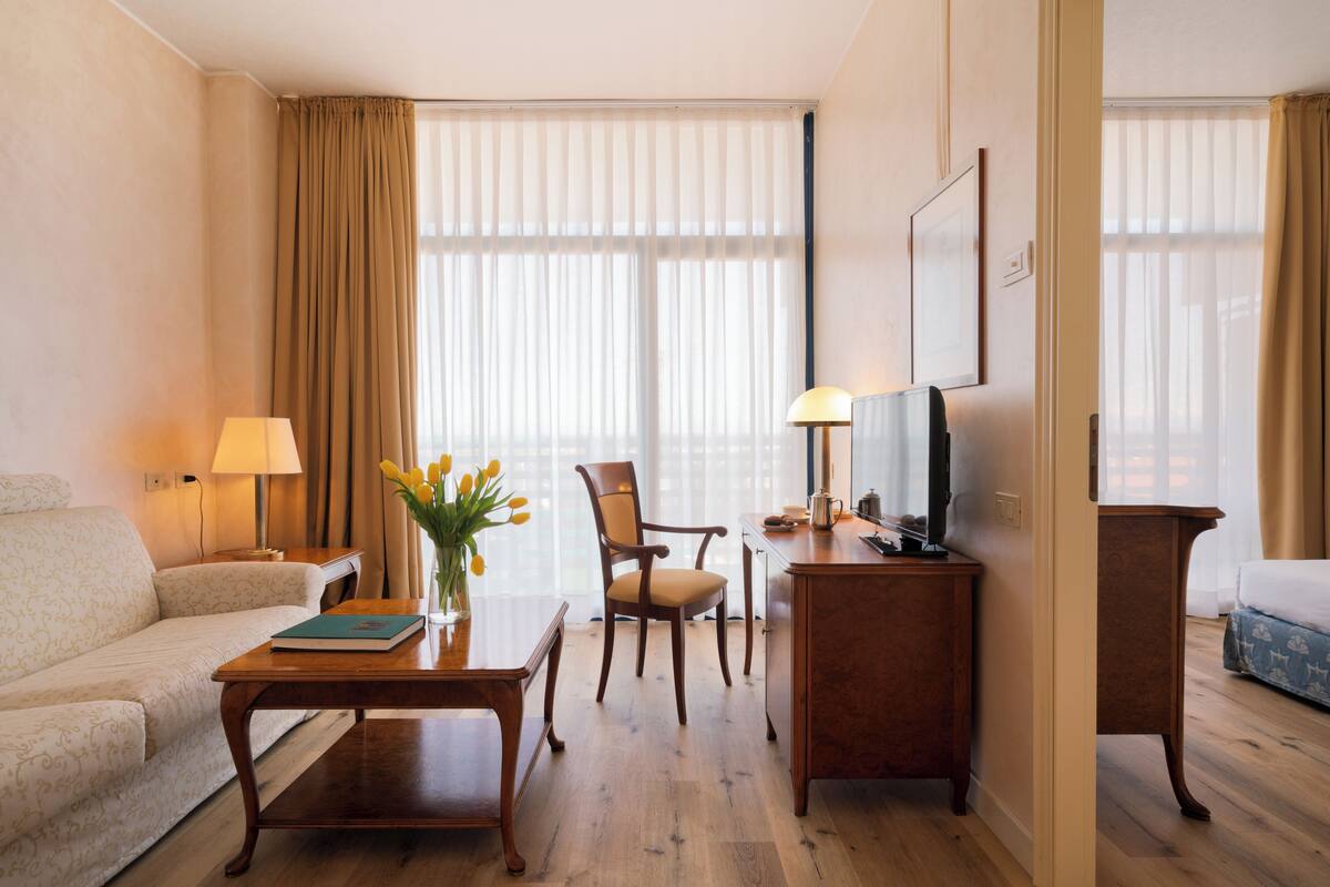 Une chambre d’hôtel avec un canapé, une table basse en bois, une chaise, un bureau avec une lampe, une télévision et une grande fenêtre avec des rideaux.