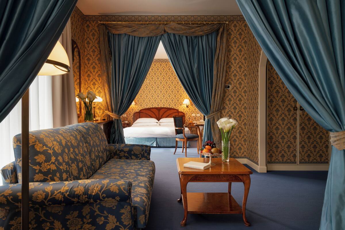 Une chambre d’hôtel avec un lit, un canapé, une table avec un livre et des fleurs, et des rideaux.
