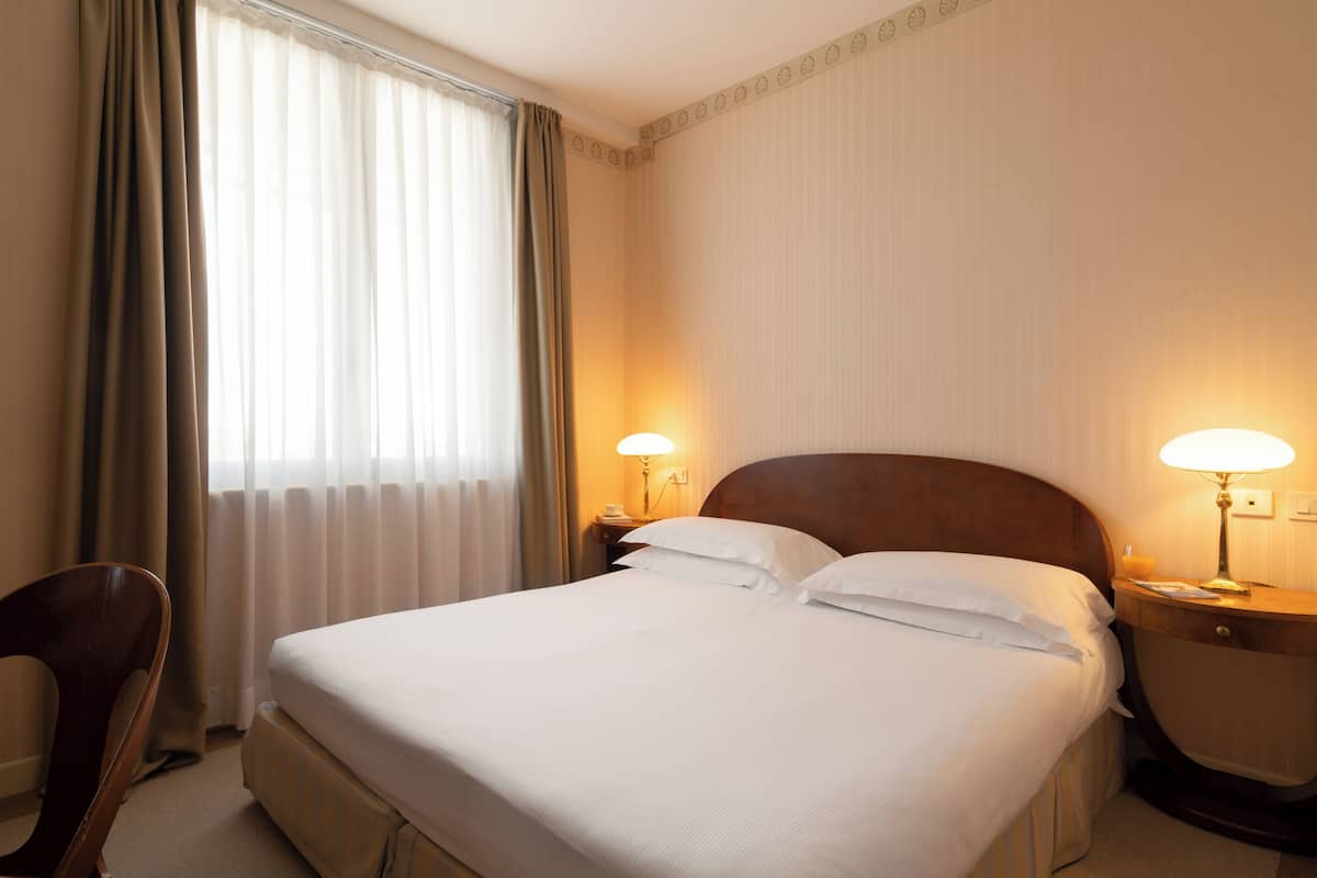 Une chambre d’hôtel avec un grand lit, deux lampes de chevet, une tête de lit en bois, une chaise et une fenêtre avec des rideaux.