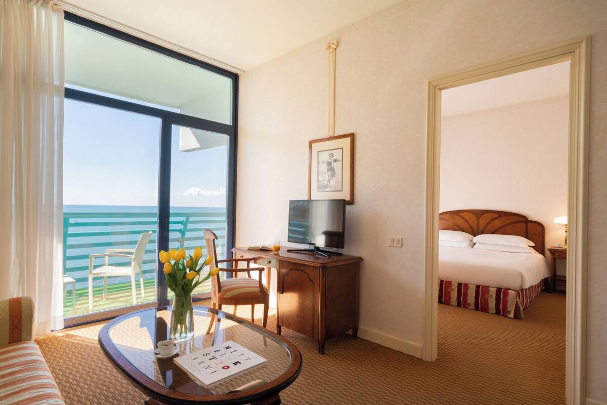 Une chambre d’hôtel avec un lit, un bureau avec un ordinateur, une chaise, une table avec un vase de fleurs et une vue sur l’océan.