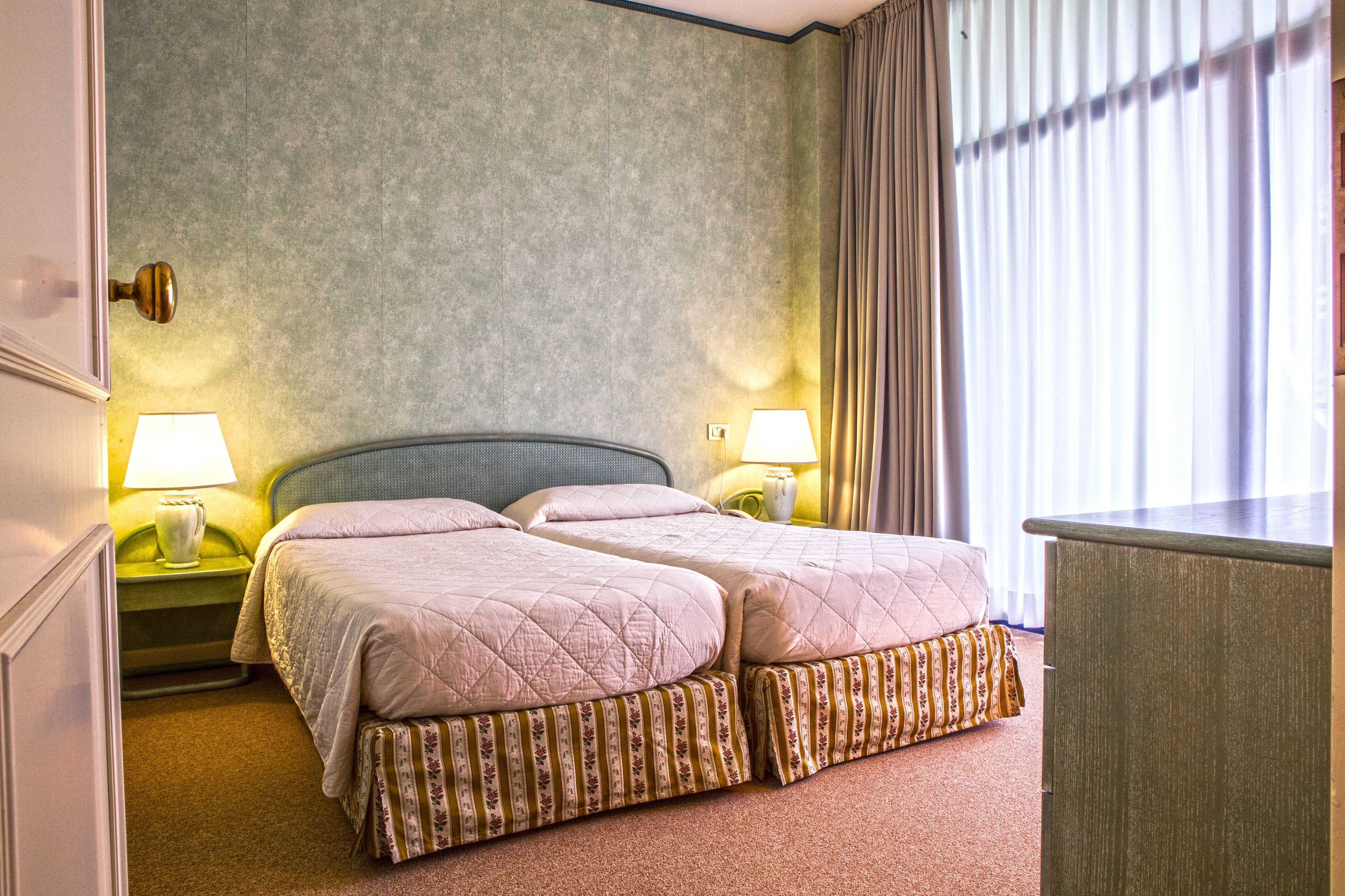 Ein Hotelzimmer mit zwei Betten, einem Nachttisch mit Lampen, einem Schreibtisch und einem Fenster mit Vorhängen.