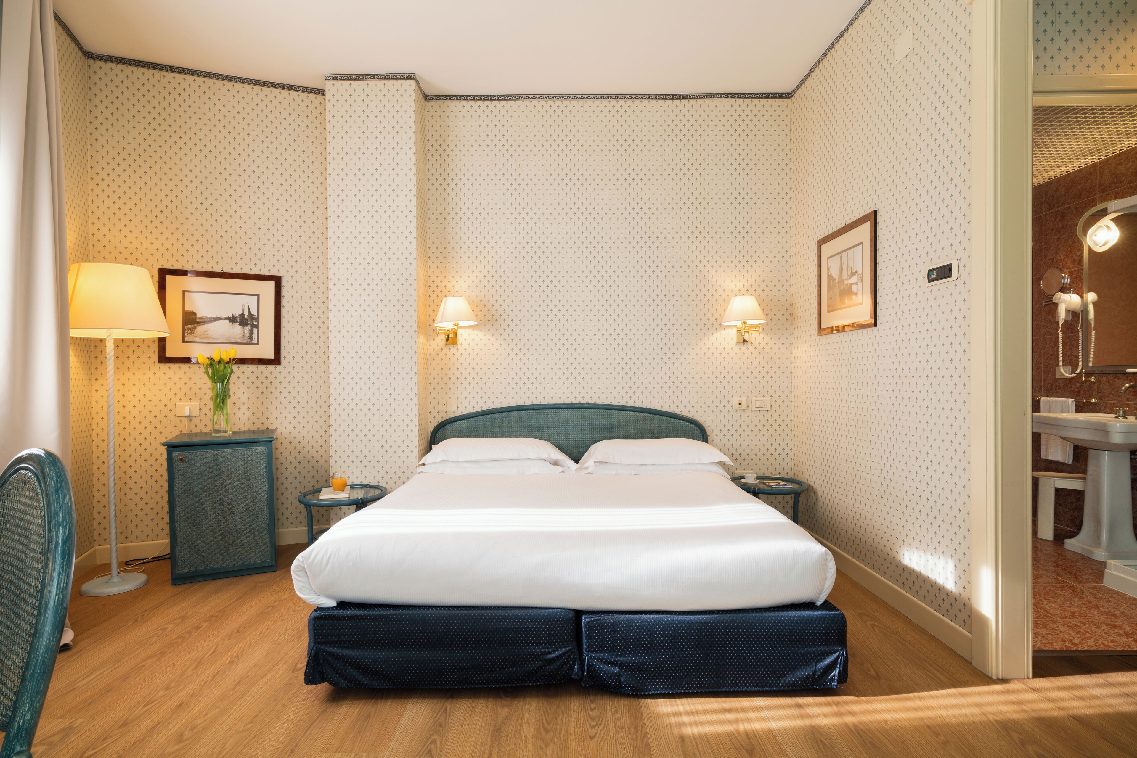 Una habitación de hotel ordenada con una cama grande, dos mesitas de noche con lámparas, una mesita auxiliar con un jarrón y una silla.