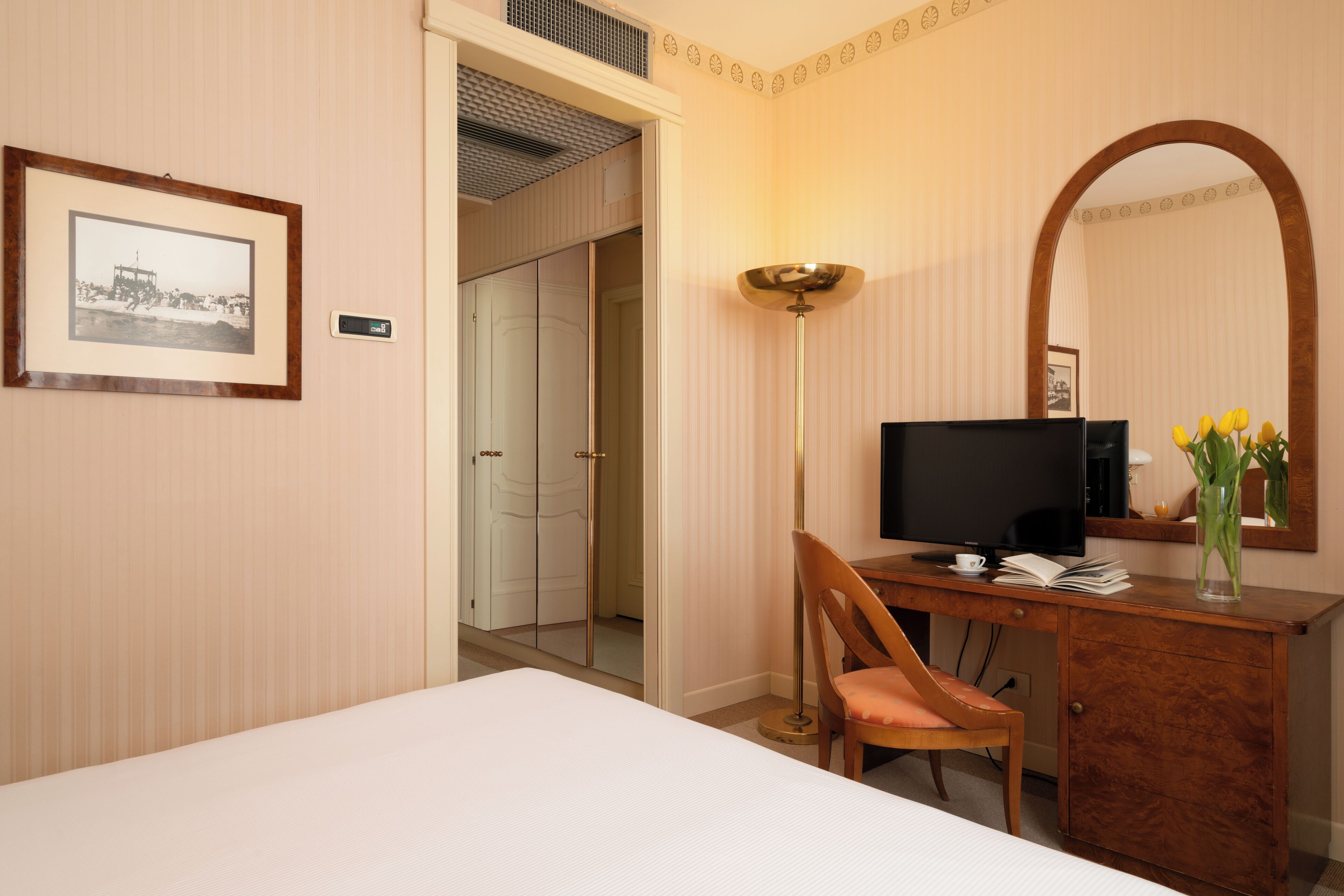 Une chambre d’hôtel comprenant un lit, un bureau avec une télévision, une chaise, une lampe de bureau, un tableau encadré au mur et un miroir.
