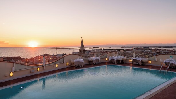A rooftop pool with tables and chairs, overlooking a sunset over the sea and a cityscape. - Grand Hotel Astoria (Grado)
