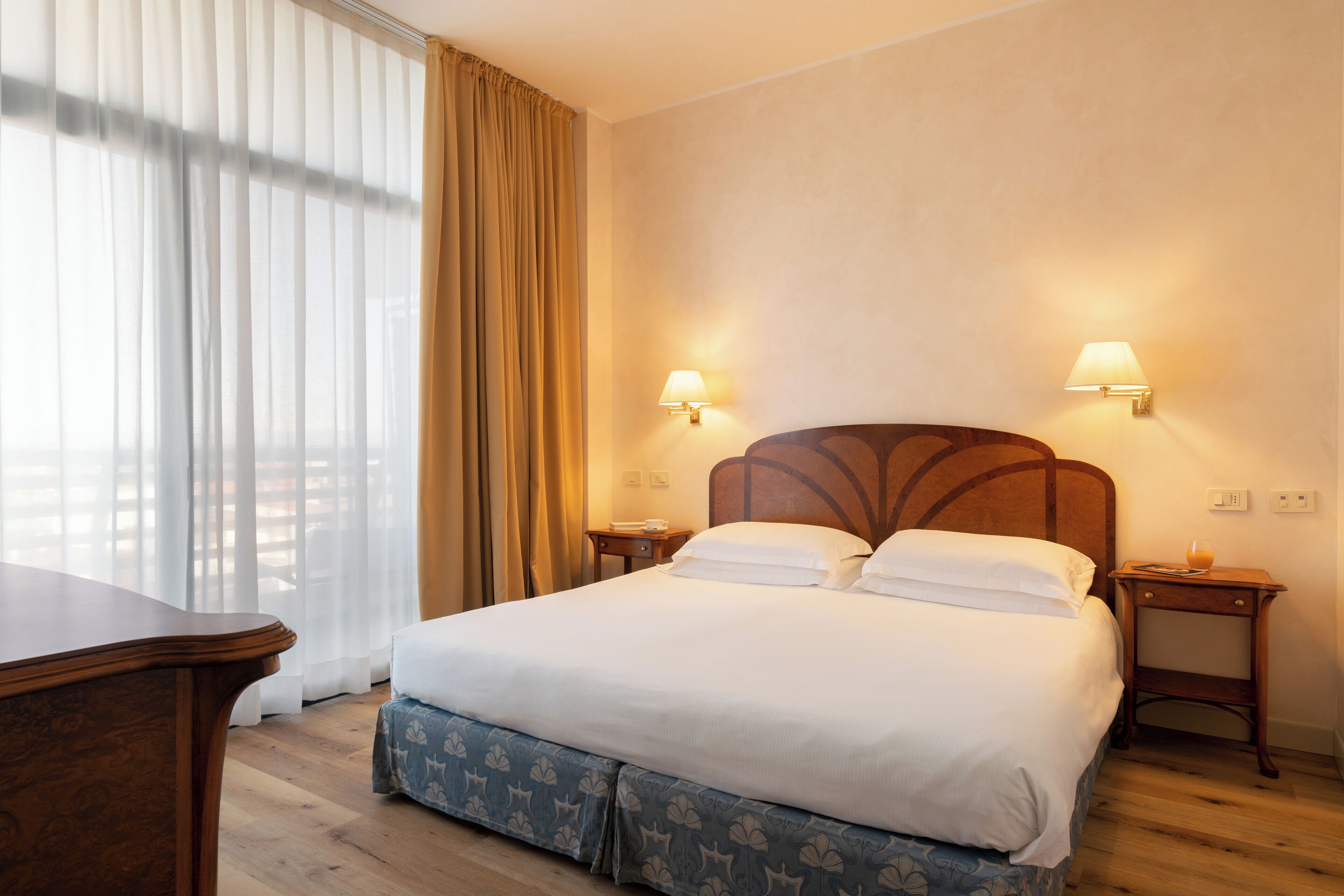 A hotel room with a bed, two bedside lamps, a wooden nightstand, a wooden desk, and a window with curtains.