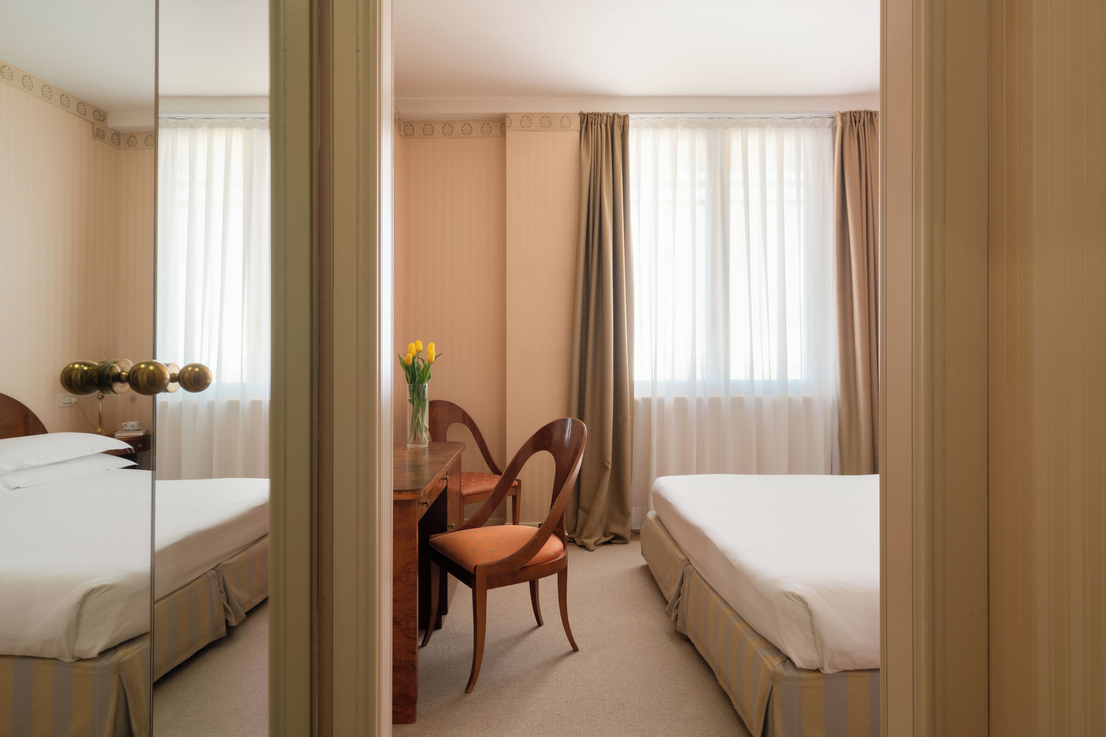 A hotel room with two beds, a wooden desk with a chair, a vase with flowers, and a large mirror.
