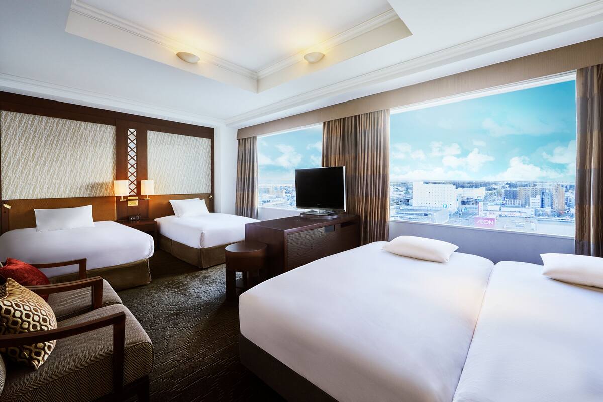 A hotel room with two beds, a desk, a television, and a large window with a city view.