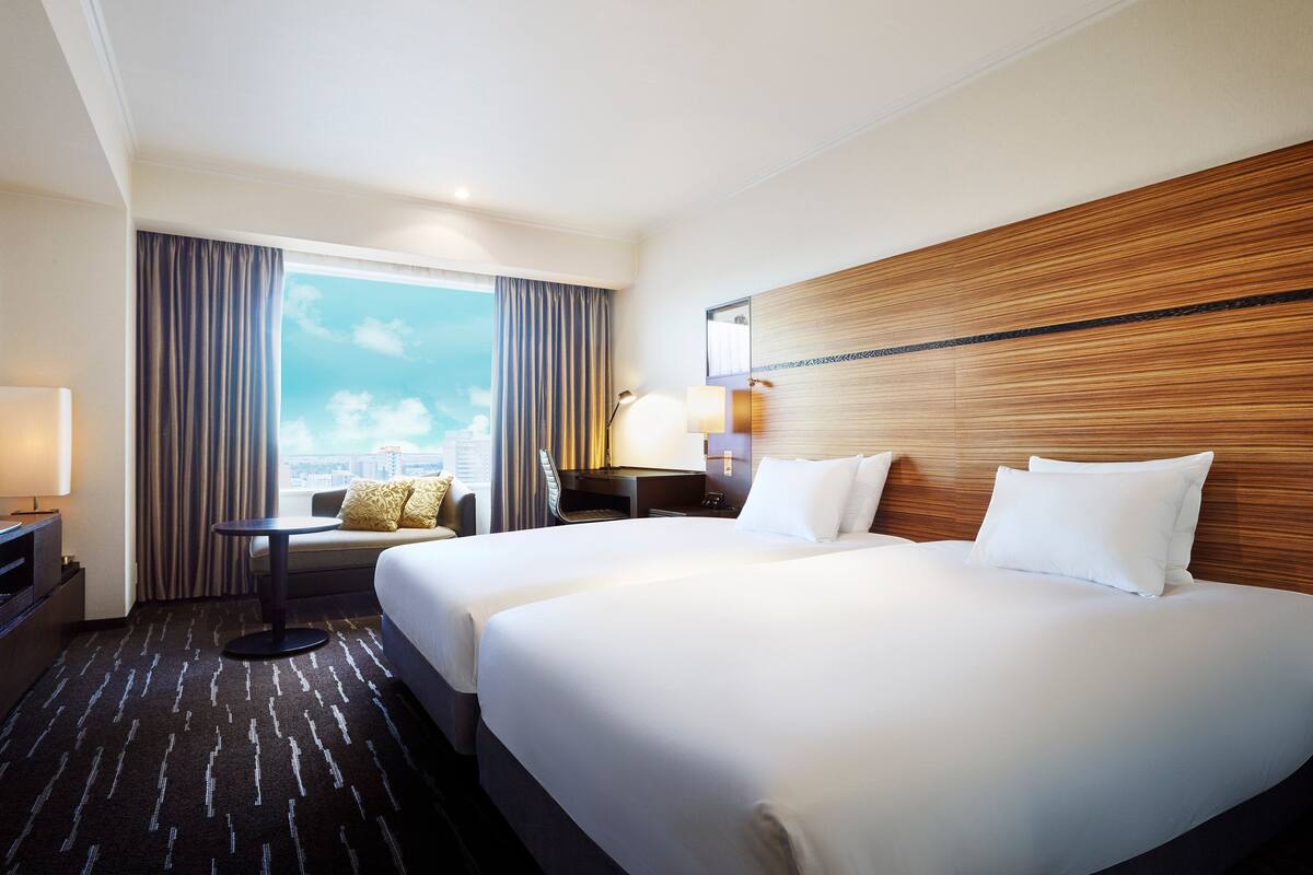 A hotel room with two beds, a desk, a chair, a window with curtains, and a city view.