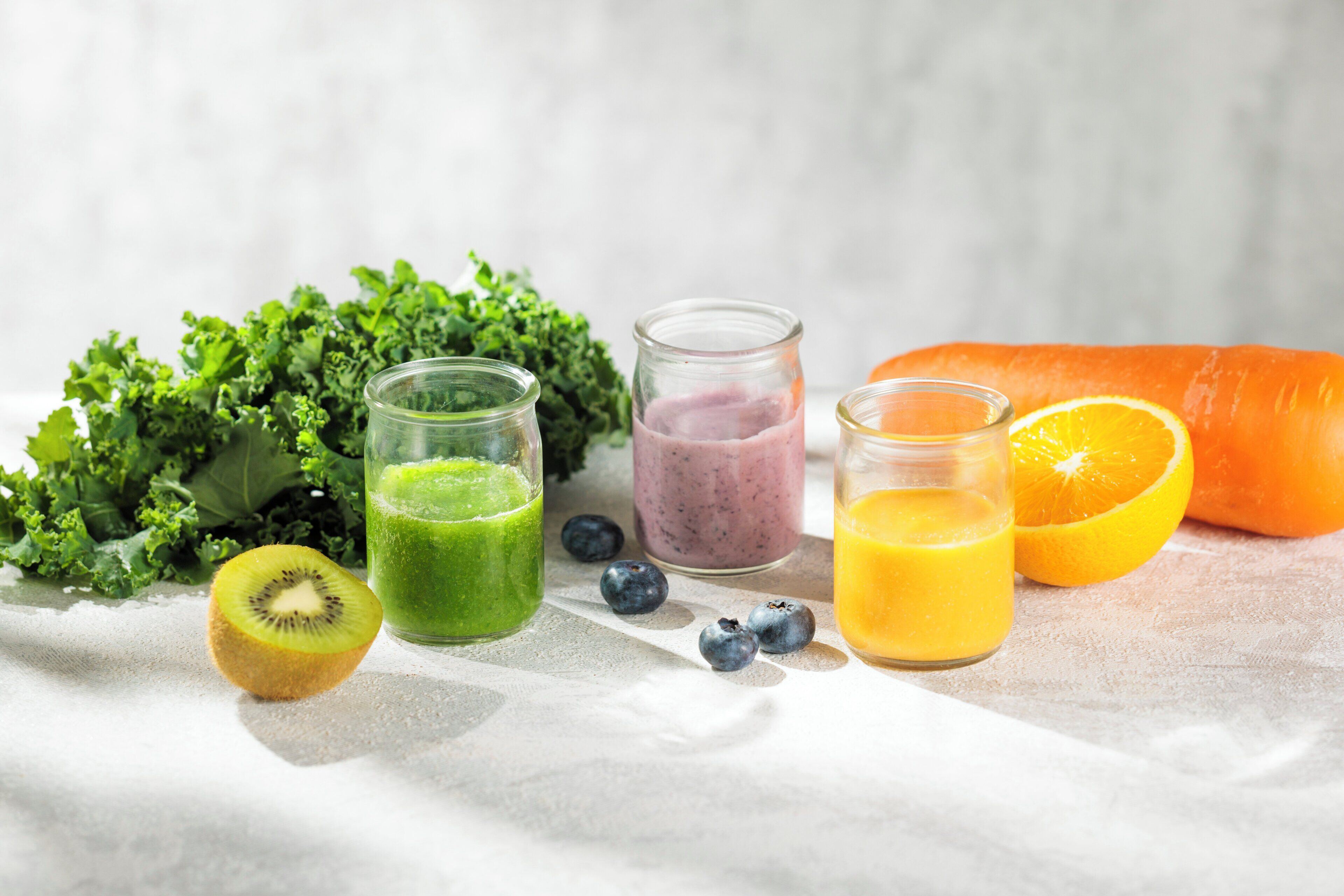A variety of fresh fruits and vegetables, including a carrot, kiwi, blueberries, and kale, alongside three different colored smoothies.