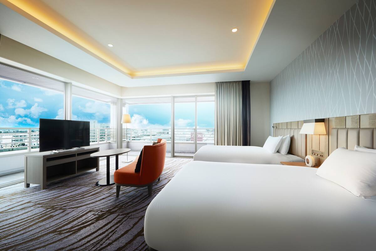 A hotel room with a large bed, a TV, a desk, and a chair. The room has a city view through large windows.