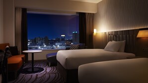 A hotel room with two beds, a small table, a chair, and a view of a cityscape at night.