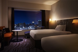 Habitación de hotel con dos camas, una mesita, una silla y vista a un paisaje urbano nocturno.