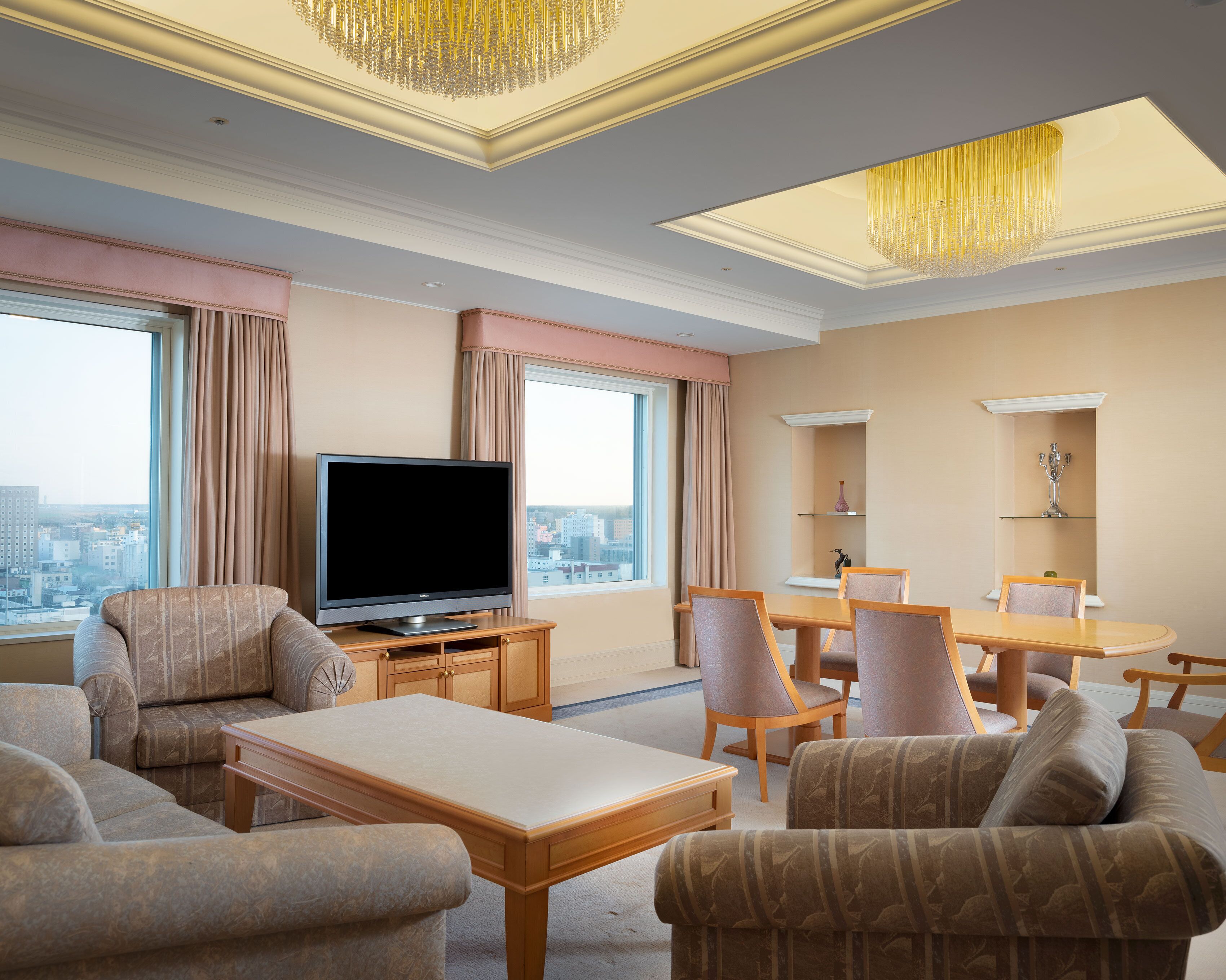 A hotel room with a large flat-screen TV, a dining table with chairs, a sofa, and a large window with curtains.
