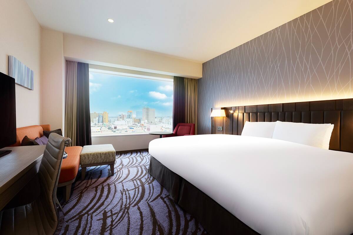 A hotel room with a large bed, a desk, a chair, a small table, and a view of the city through a large window.