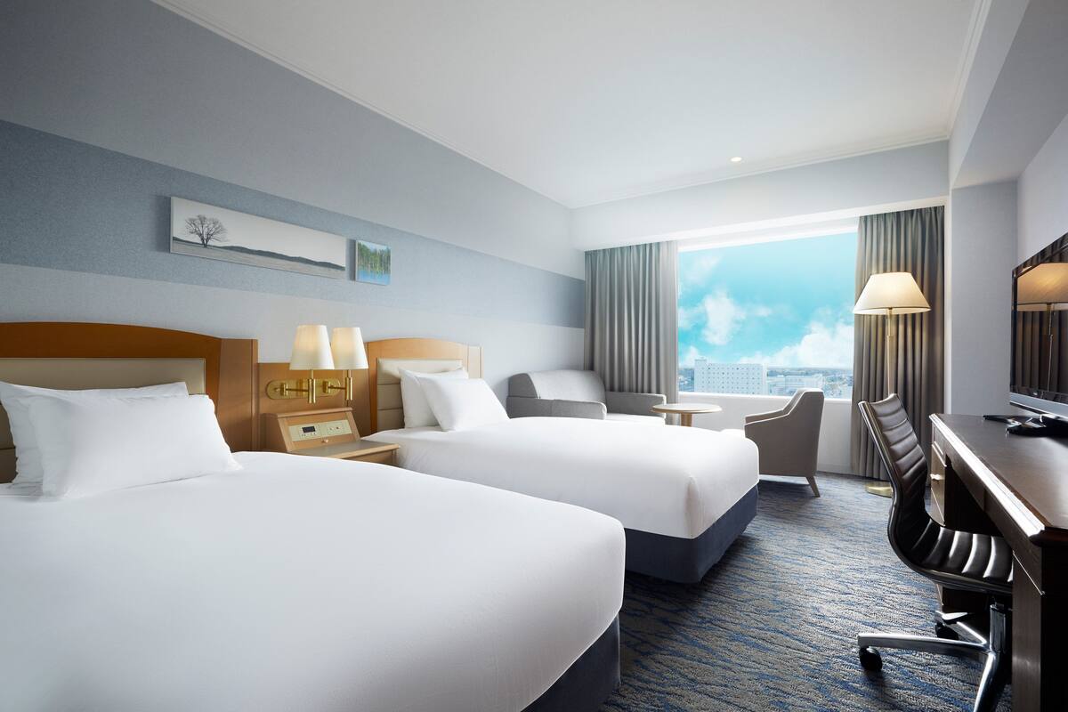 A hotel room with two beds, a desk, a chair, a TV, and a view of the city.