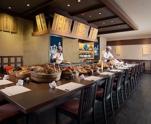 A restaurant with a long dining table, multiple serving stations, and a sushi bar.