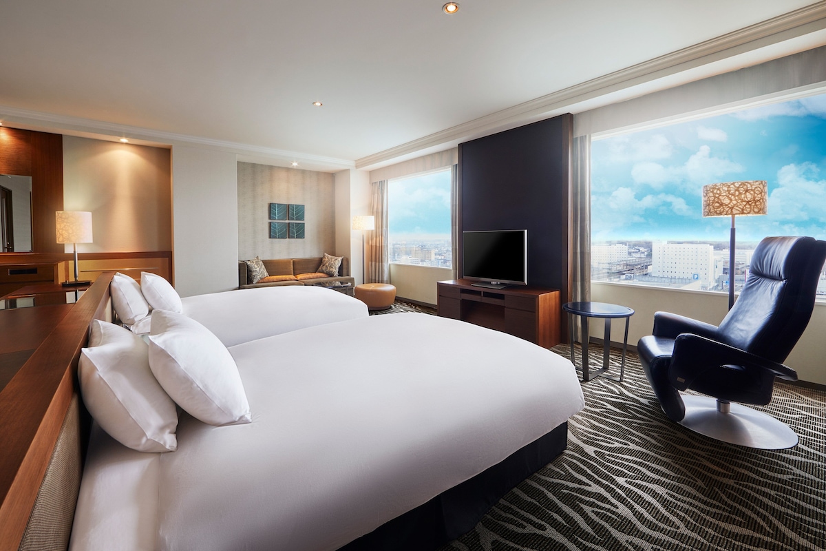 Habitación de hotel con una cama grande, un televisor de pantalla plana, una zona de estar con una silla y una mesa, y vistas a la ciudad desde la ventana.
