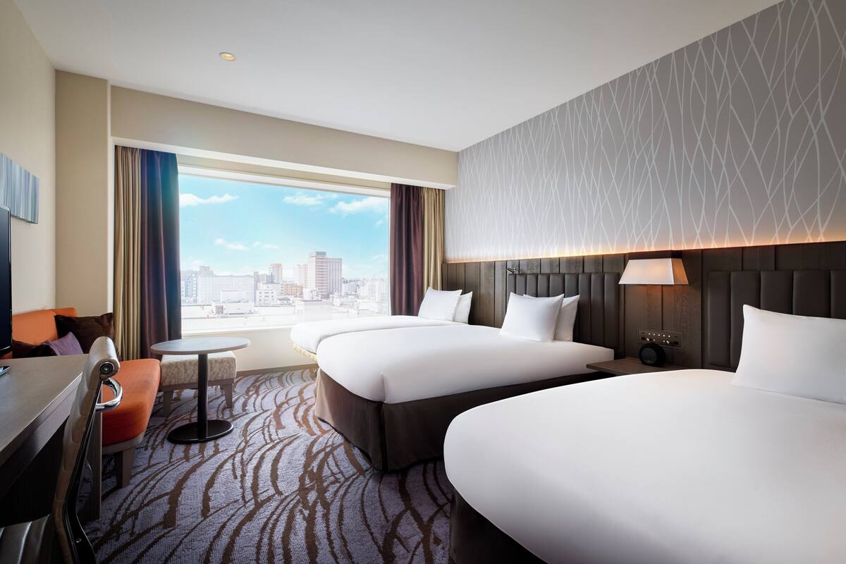 A hotel room with two beds, a desk, a chair, a small table, a lamp, and a view of the city.