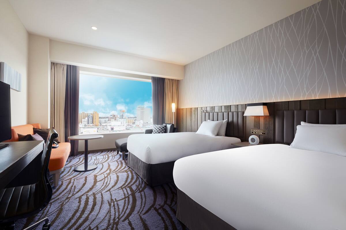 A hotel room with two beds, a desk, a chair, a small table, and a view of the city.