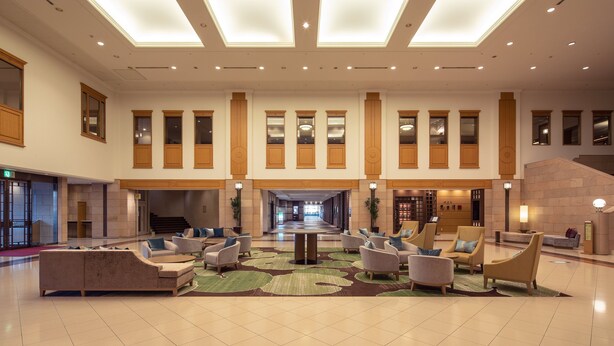 A spacious lobby with a high ceiling, large windows, and a central seating area with multiple chairs and a table.