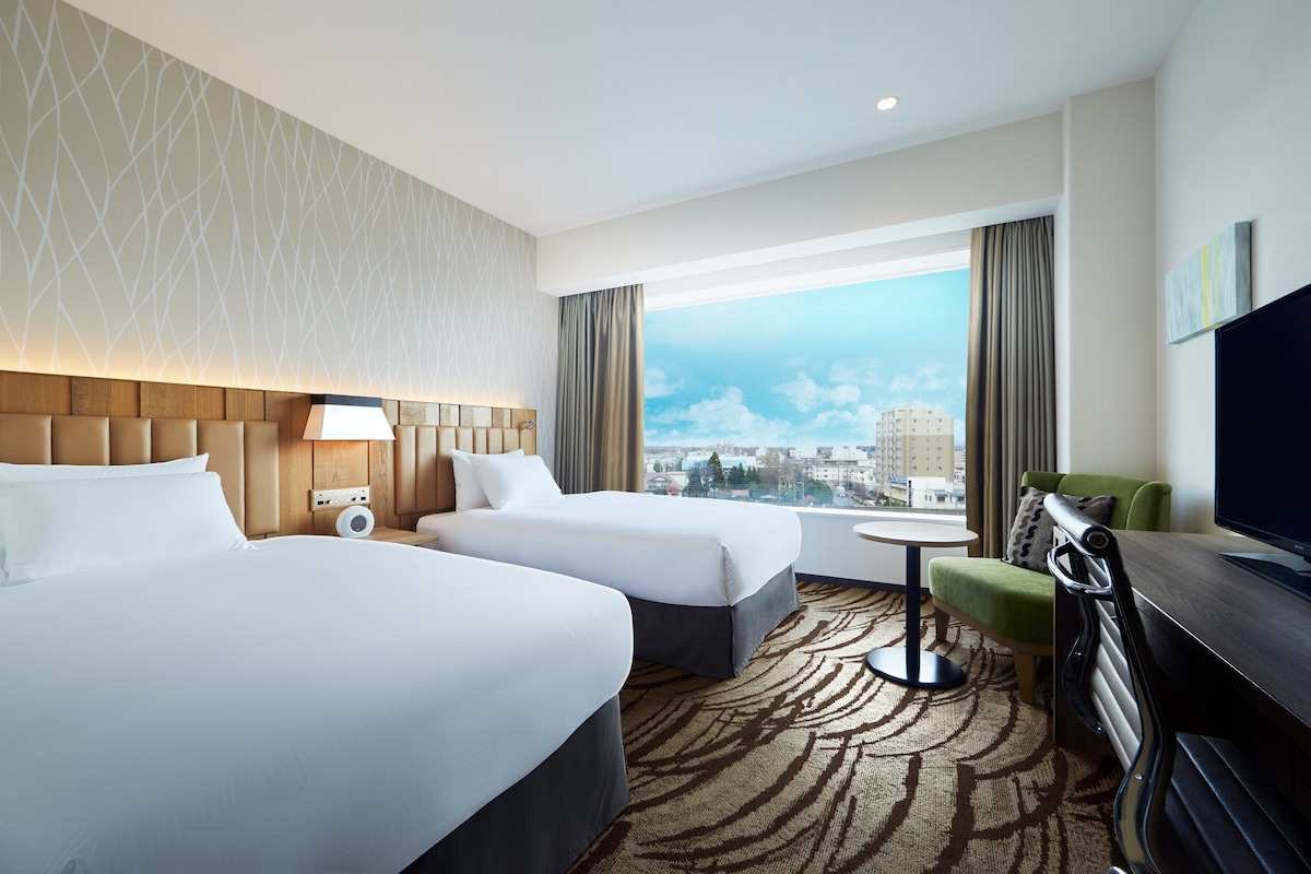 Habitación de hotel con dos camas, televisión, escritorio y vistas a la ciudad.