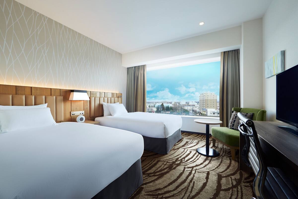 A hotel room with two beds, a TV, a desk, and a view of the city.