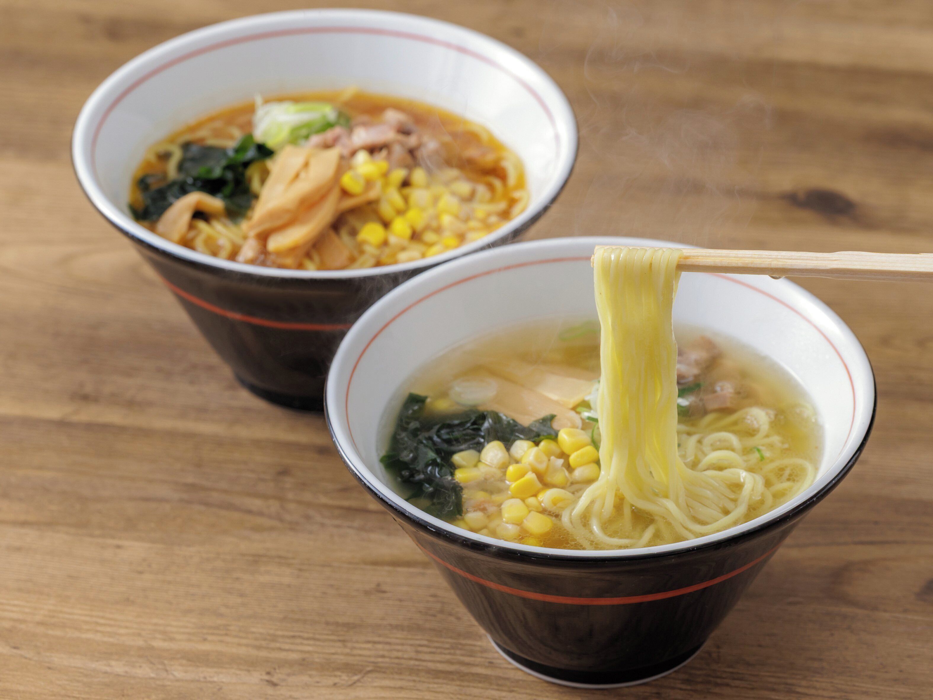 Two bowls of soup with noodles and vegetables on a wooden table.