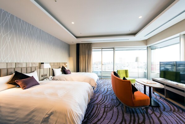 A hotel room with two beds, a desk, a chair, a TV, and a large window with a city view.