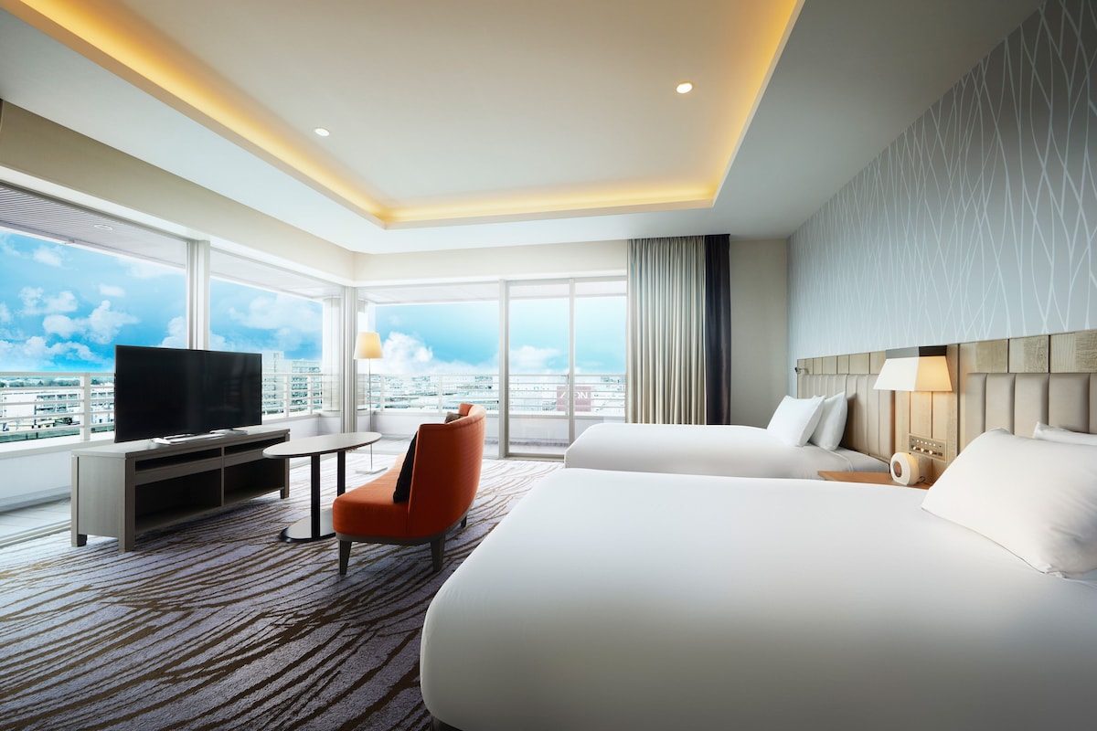 Habitación de hotel con una cama grande, televisión, escritorio y silla. Ventanas amplias ofrecen vista a la ciudad.