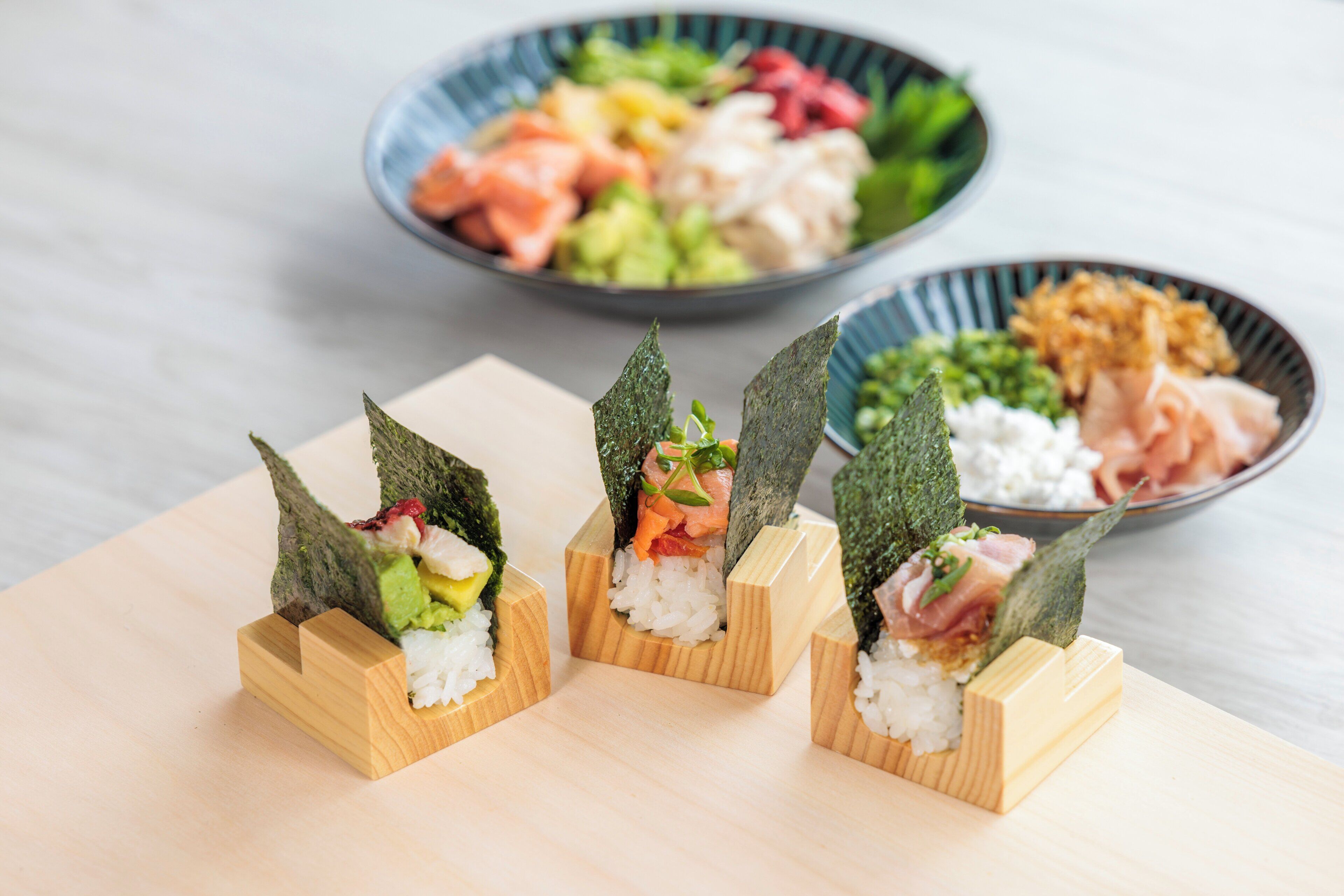 Three sushi rolls on wooden blocks with seaweed on top, a bowl of assorted sashimi in the background, and a bowl of pickled vegetables.