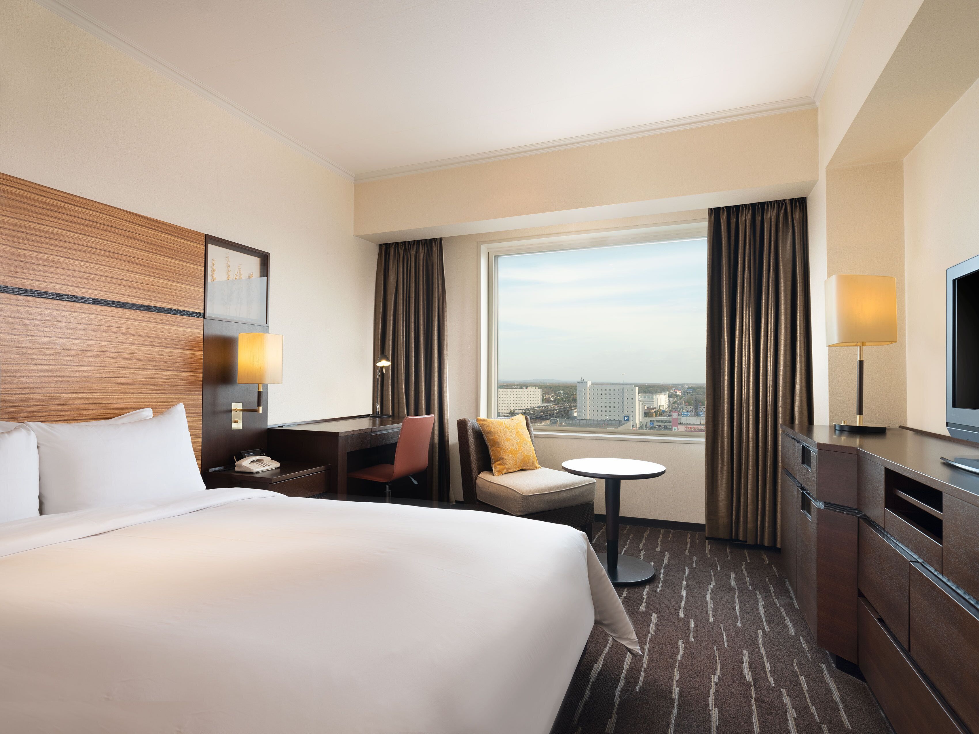 A hotel room with a large bed, a desk, a chair, a small table, a TV, and a window with a city view.